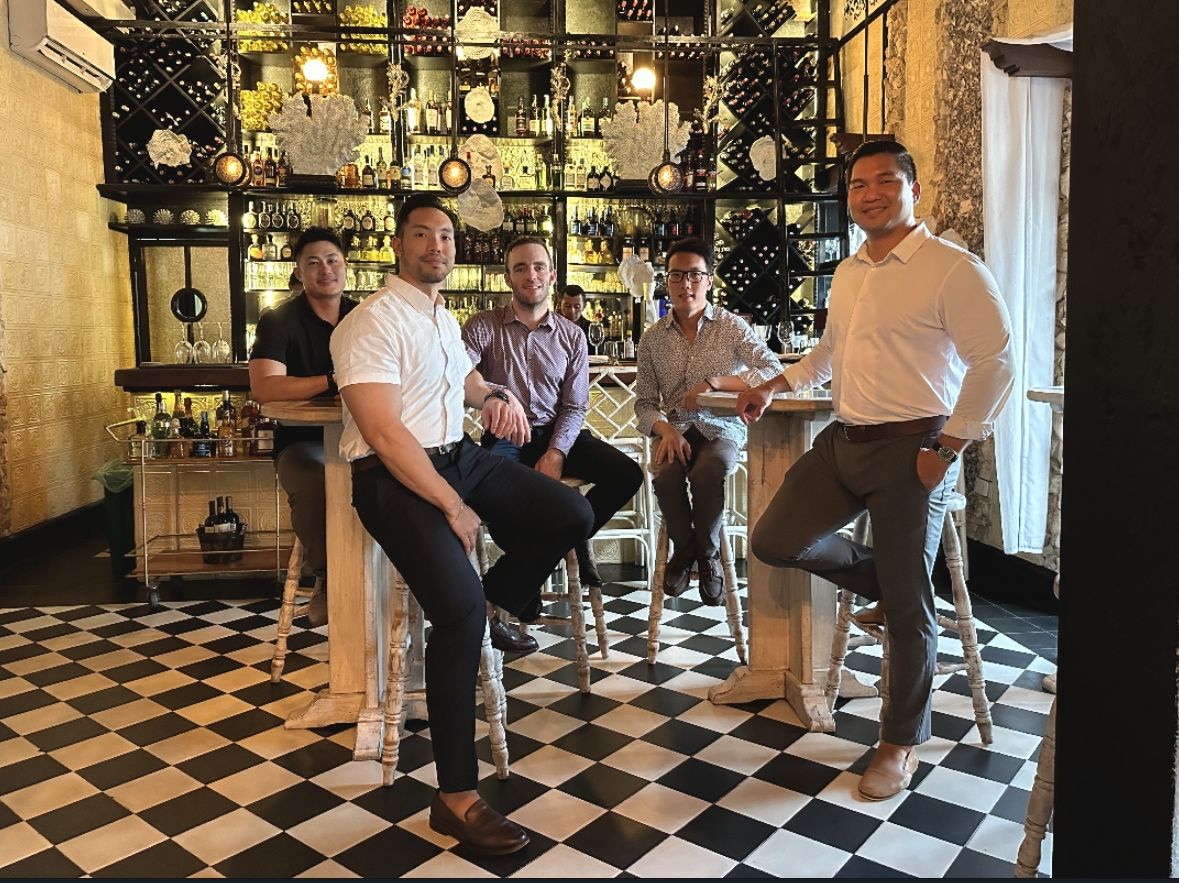 Five friends posed around the counter of a wood-panelled wine bar, David standing at the right.